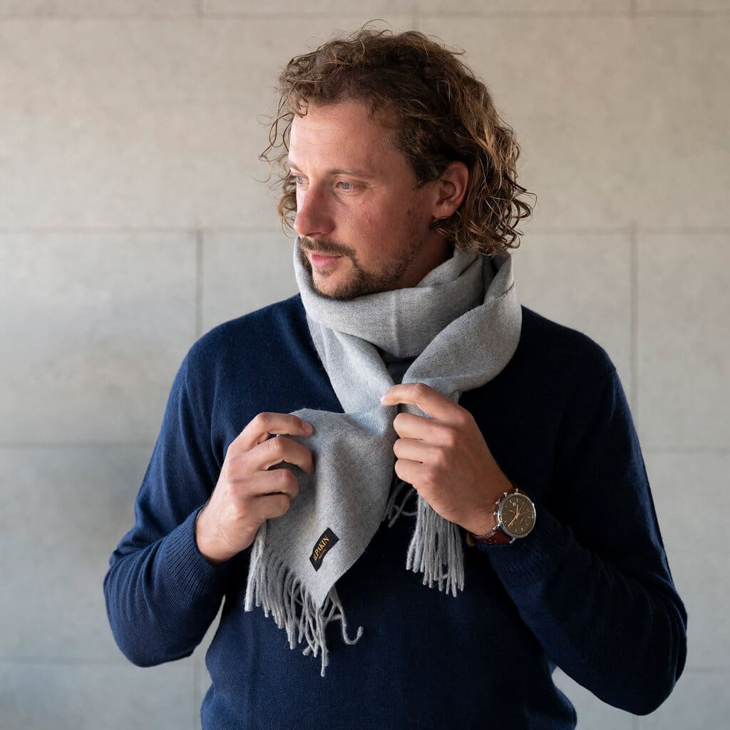 Mann mit Locken trägt einen Naturfarbe Alpaka Schal in hellgrau und einen dunkelblauen Pullover