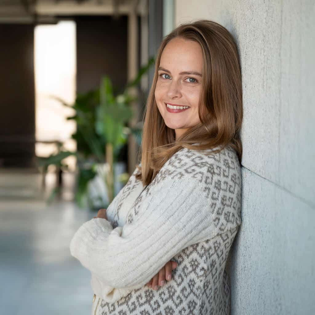 Frau mit verschränkten Armen lehnt an einer hellen Wand und trägt ihre warme Alpaka Strickjacke in creme