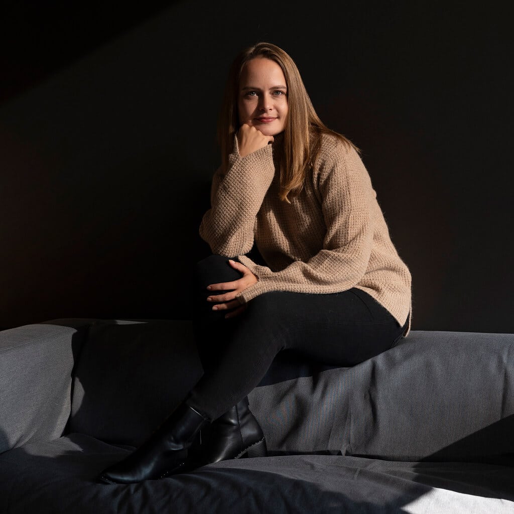 Alpaka Pullover Nuna in Naturfarbe hellbraun, getragen von blondem Model, das auf einem dunklem Sofa sitzt