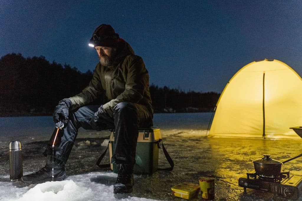 Mann auf Winterexpedition beim Eisangeln mit beleuchtetem Zelt im Hintergrund